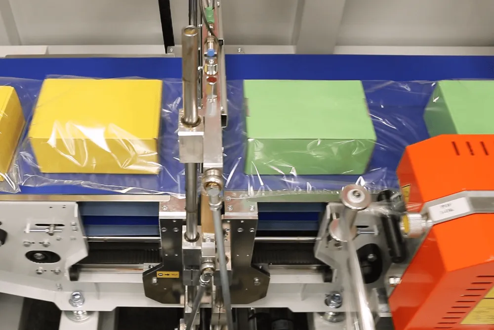 Boxes riding a conveyor inside a continuous tube of clear film, with the cross seal bar above and the orange side seal unit at the right edge.