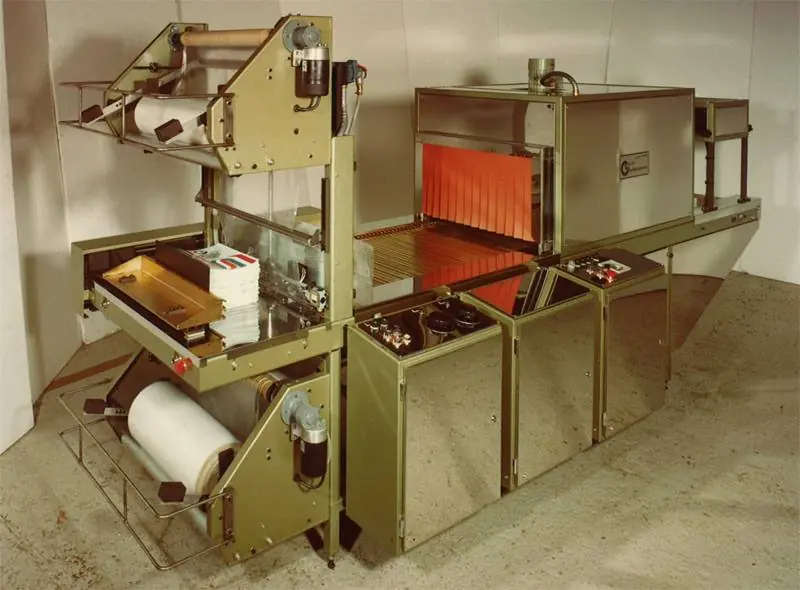 An early L-sealer with a roll of film on the left, the L-shaped seal bar in the middle, and a shrink tunnel with orange curtains on the right.