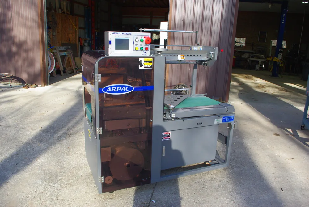 A gray Arpac automatic L-bar sealer on casters with a touchscreen control box on top, a tinted guard door on the left, and a green conveyor belt on the right.