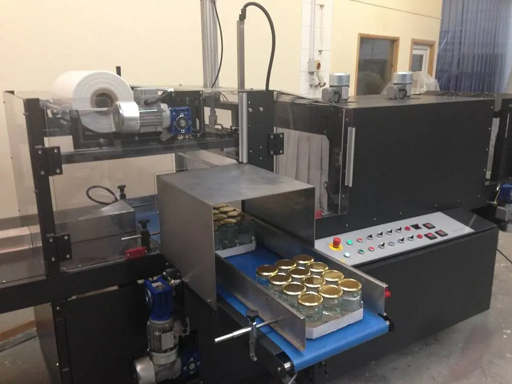 A shrink wrapping machine with a film roll on top and a blue conveyor carrying glass jars through it.