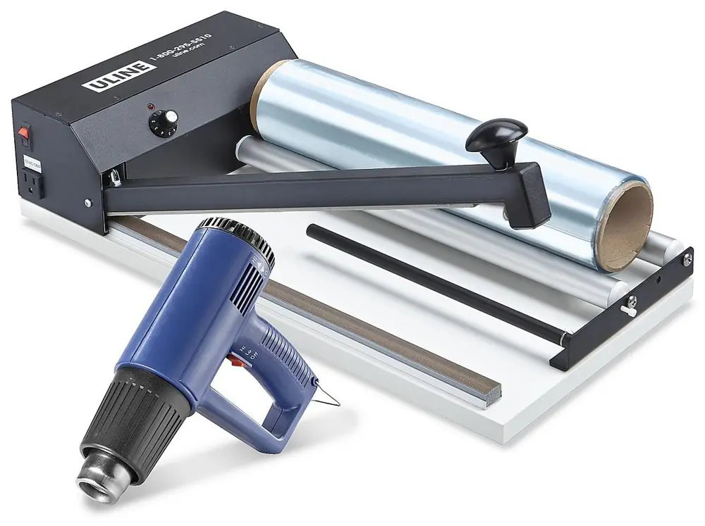 A tabletop I-bar sealer with a roll of shrink film behind the bar and a blue heat gun in front.